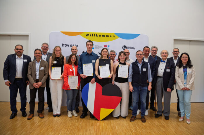Gruppenfoto der Verleihung des Helmut Schmidt – Valéry Giscard d’Estaing-Preises 2026 am Deutsch-Französischen Hochschulinstitut (DFHI): Die ausgezeichneten Studierenden stehen gemeinsam mit Vertreterinnen und Vertretern des DFHI-Fördervereins, der htw saar und der Université de Lorraine vor einer Veranstaltungswand mit den Logos der Hochschulen. Einige der Preisträgerinnen und Preisträger halten ihre Urkunden in den Händen. Im Vordergrund steht ein großes Herz in den Farben der deutsch-französischen Flaggen.